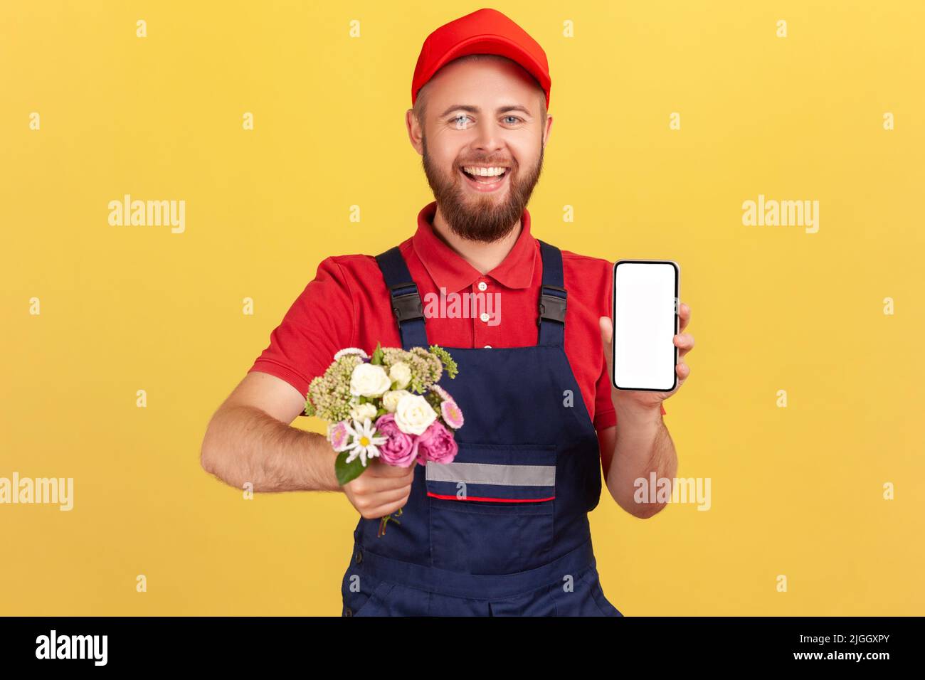 Lächelnder, positiver Mann, Kurier, der mit Blumen in den Händen stand und das Handy mit leerem Bildschirm für Werbung zeigte und auf die Kamera schaute. Innenaufnahme des Studios isoliert auf gelbem Hintergrund. Stockfoto