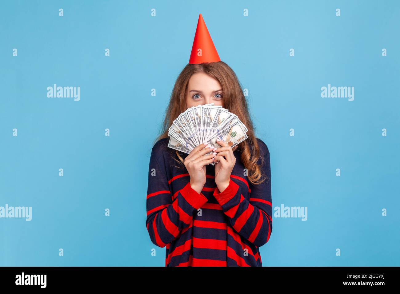 Schüchterne Frau mit gestreiftem Pullover im lässigen Stil und rotem Partykegel, die sich hinter Geldscheinen versteckt, genießt das reiche Leben und ist mit dem Geldgeschenk zufrieden. Innenaufnahme des Studios isoliert auf blauem Hintergrund. Stockfoto