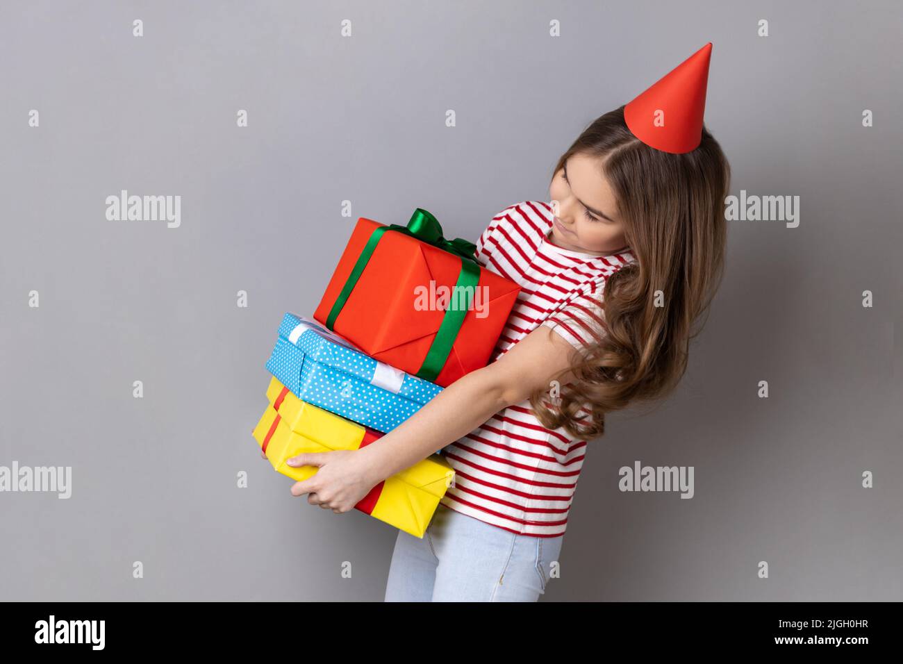 Seitenansicht Porträt eines kleinen Mädchens, das gestreiftes T-Shirt trägt und im Partykegel einen schweren Stapel von Geschenken hält, um ihren Geburtstag zu feiern. Innenaufnahme des Studios isoliert auf grauem Hintergrund. Stockfoto
