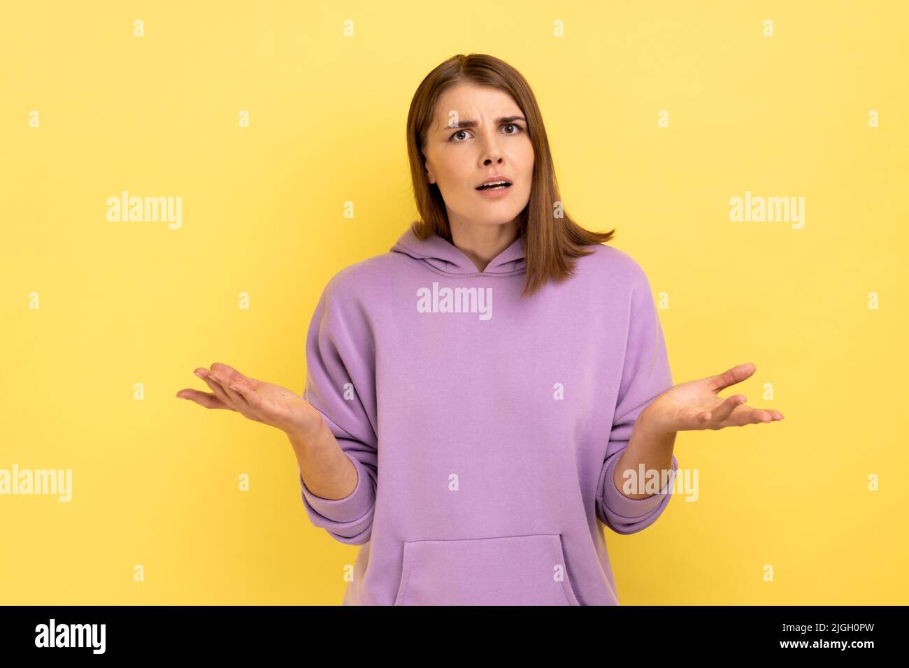 Porträt einer unglücklich gestressten jungen erwachsenen Frau, die mit erhobenem Arm steht und fragt, negative Emotionen ausdrückt und einen violetten Hoodie trägt. Innenaufnahme des Studios isoliert auf gelbem Hintergrund. Stockfoto