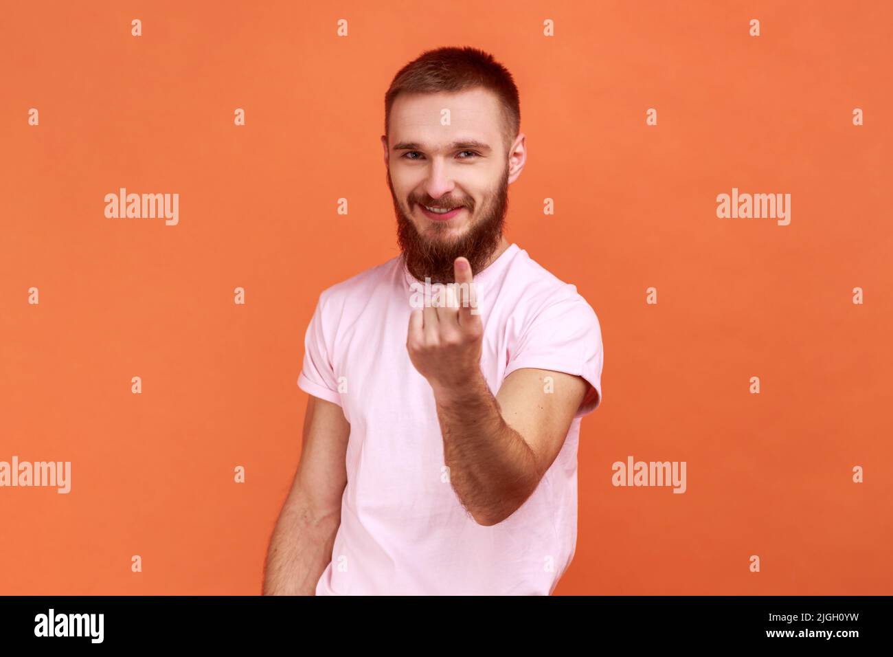 Porträt eines bärtigen Mannes, der Sie mit dem Finger anruft, mit spielerischem Ausdruck auf die Kamera schaut, beim Date flirtet und einladend, in einem rosa T-Shirt. Innenaufnahme des Studios isoliert auf orangefarbenem Hintergrund. Stockfoto