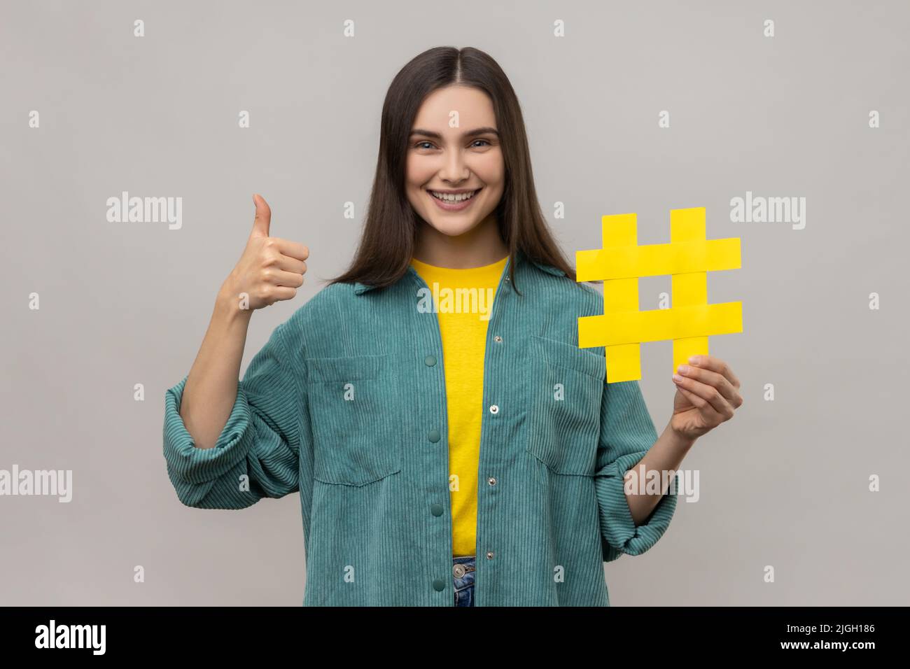Wie und folgen Sie Trends, beliebte Blog. Optimistische Frau mit Hashtag-Symbol für soziale Medien und Daumen nach oben Geste in lässiger Jacke. Innenaufnahme des Studios isoliert auf grauem Hintergrund. Stockfoto