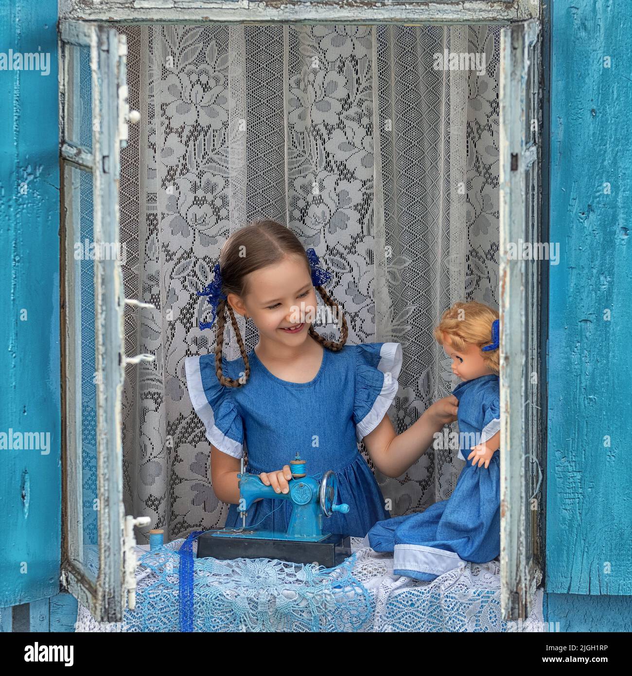 Porträt eines Schneidermädchen Nähen Sie Puppenkleidung auf einer Kinder-Nähmaschine im Fenster eines alten Holzhauses. Stockfoto