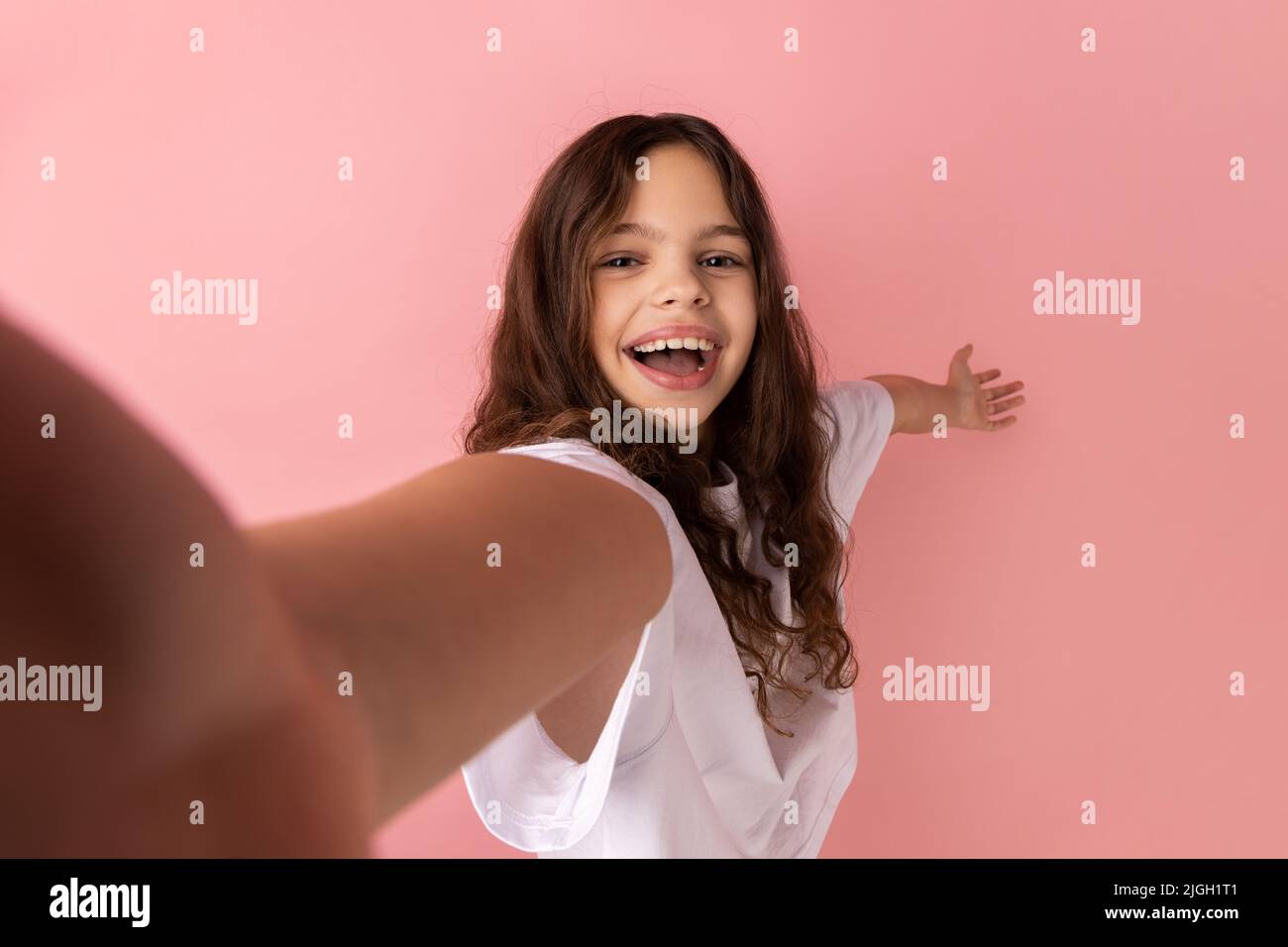 Porträt eines kleinen Mädchens mit weißem T-Shirt, das Selfie macht, auf den POV der Kamera schaut und eine willkommene Geste zeigt, aus der Perspektive des Fotos. Innenaufnahme des Studios isoliert auf rosa Hintergrund. Stockfoto