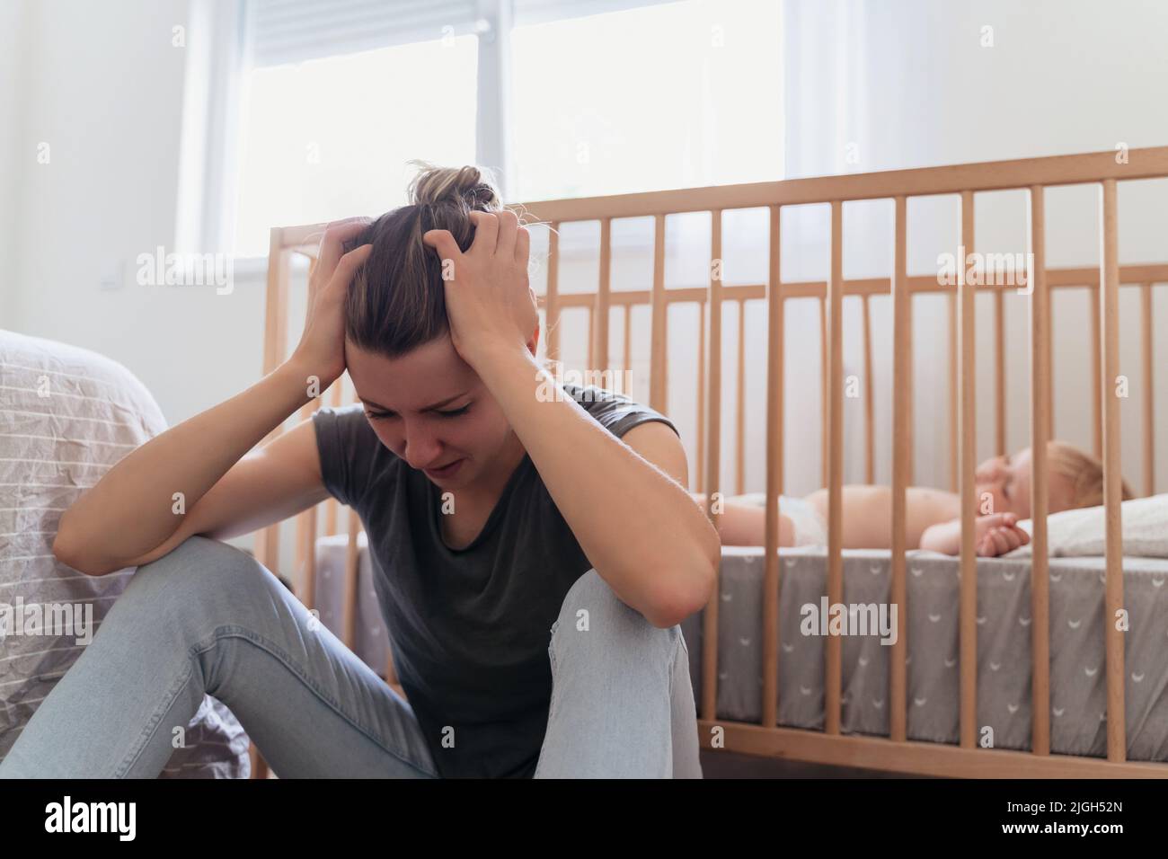 Die junge Mutter hält den Kopf in den Händen vor Verzweiflung, fühlt sich müde und erschöpft, weint neben dem Baby, das im Bett schläft, leidet an einer postnatalen Depression, die einer psychologischen Behandlung bedarf Stockfoto