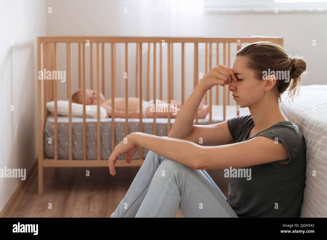 Seitenansicht einer unglücklich frustrierten jungen Mutter, die im Kinderschlafzimmer auf dem Boden sitzt, während das Baby im Bett schläft und versucht, sich zu beruhigen, und an postnatalen Depressionen leidet Stockfoto