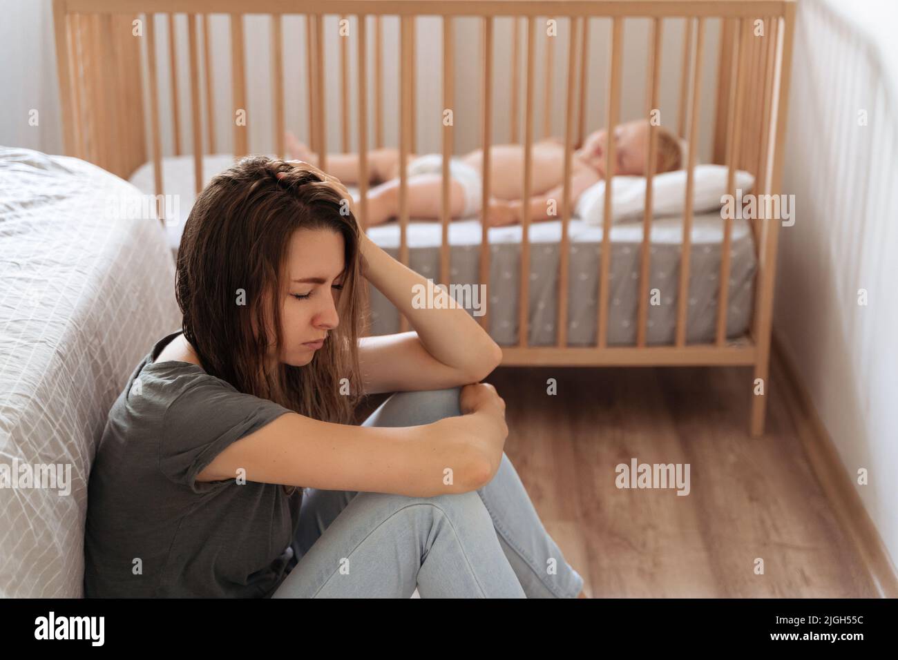 Junge, müde Mutter, die an postnataler Depression leidet, sitzt auf dem Boden neben dem schlafenden Baby in der Krippe mit geschlossenen Augen. Fühlen Sie sich erschöpft depressiv und fordern psychologische Hilfe und Unterstützung Stockfoto
