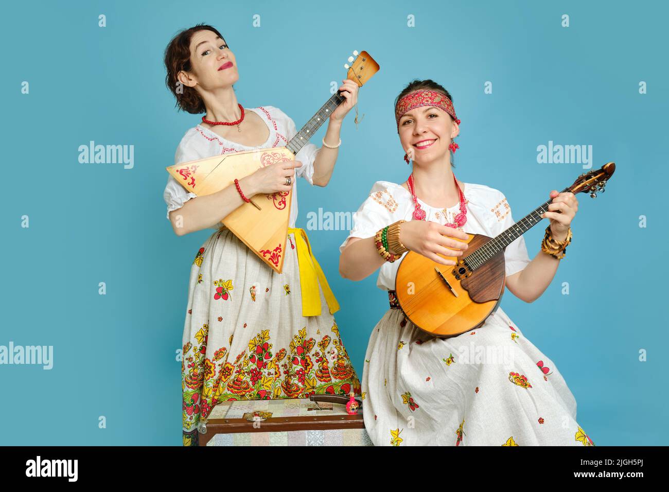 Musikerinnen in russischen Volkskleidern mit Musikinstrumenten auf blauem Studiohintergrund. Glückliche Künstler aus Russland in weißer Nationalkleidung mit Stockfoto