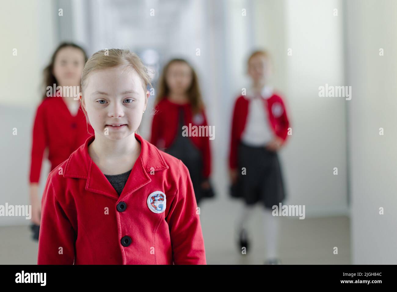 Fröhliche Schulkameradinnen mit Down-Syndrom in Uniform, die im Schulkorridor mit Klassenkameraden hinter ihr spazieren gehen. Stockfoto