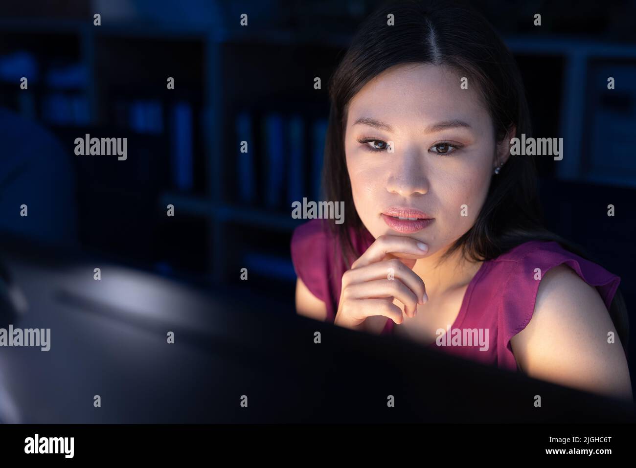 Nachdenkliche asiatische Profikerin mit Hand am Kinn, die nachts im Büro auf den Computerbildschirm schaut Stockfoto
