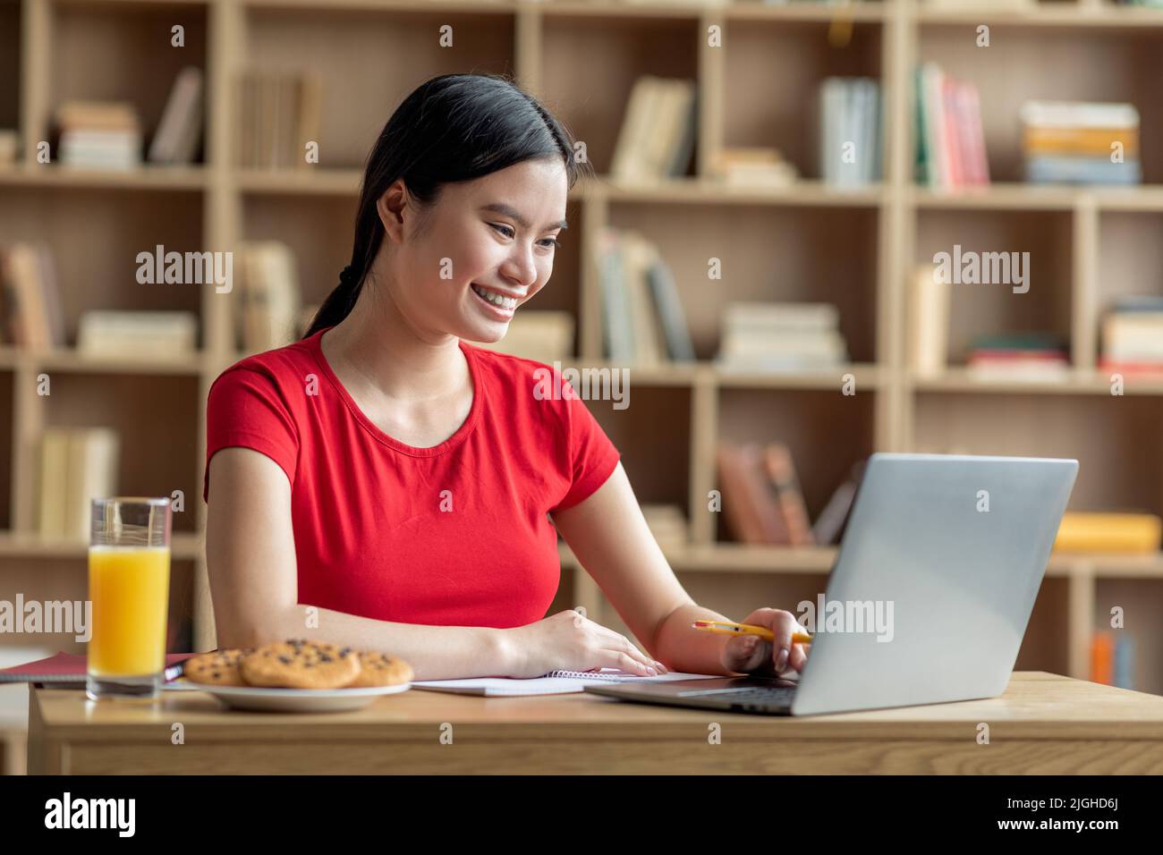 Fröhliche junge asiatische Dame Student beobachten Online-Unterricht auf Laptop, chatten und frühstücken Stockfoto