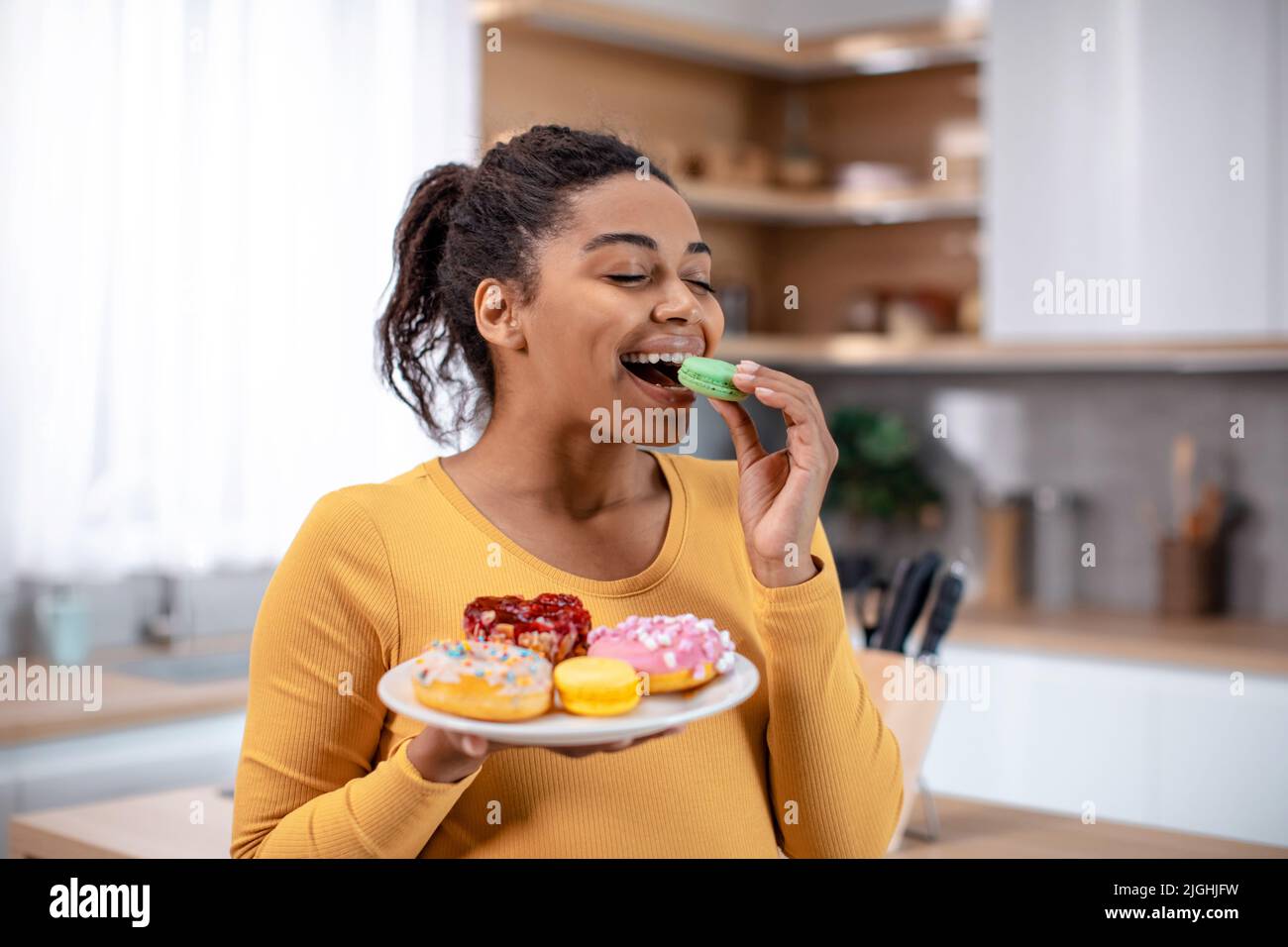 Frohes Millennial schwanger afroamerikanische Frau halten Teller mit Süßigkeiten und essen Cookies Stockfoto