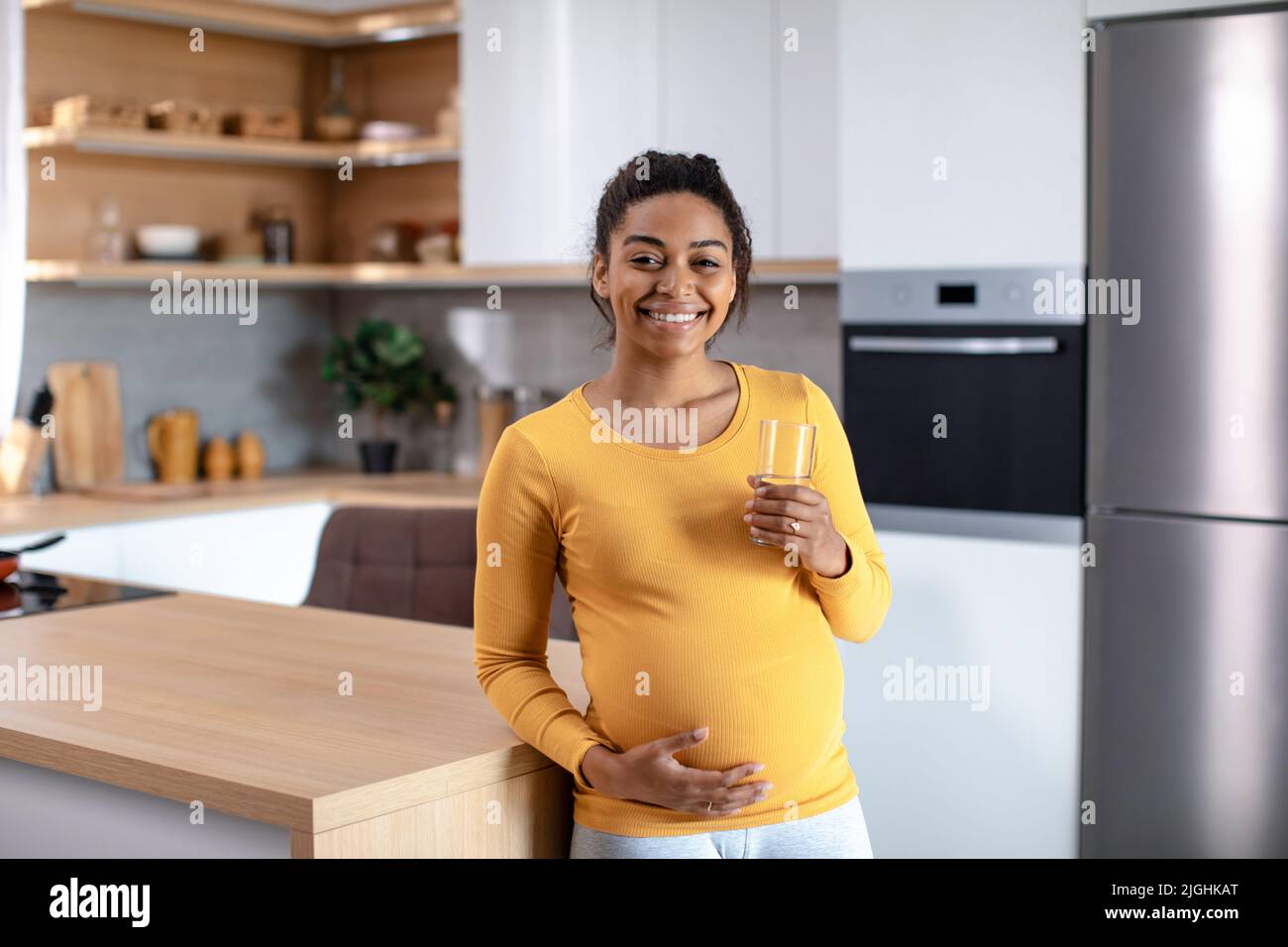 Glückliche junge, schwanger afroamerikanische Frau, die großen Bauch berührt und ein Glas Wasser hält Stockfoto