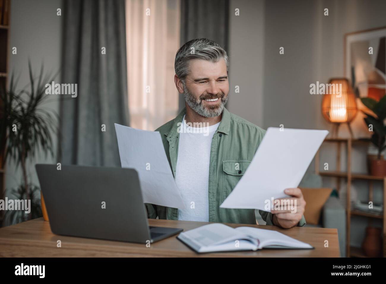 Lächelnder kaukasischer älterer Mann mit Bart arbeitet auf einem Laptop mit Dokumenten im Wohnzimmer Stockfoto