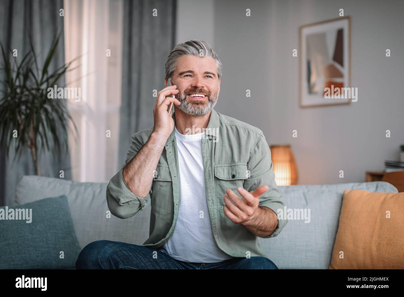 Fröhlicher, älterer kaukasischer Mann mit Bartrufen im Wohnzimmer. Im Ruhestand im Gespräch mit dem Kunden zu Hause Stockfoto