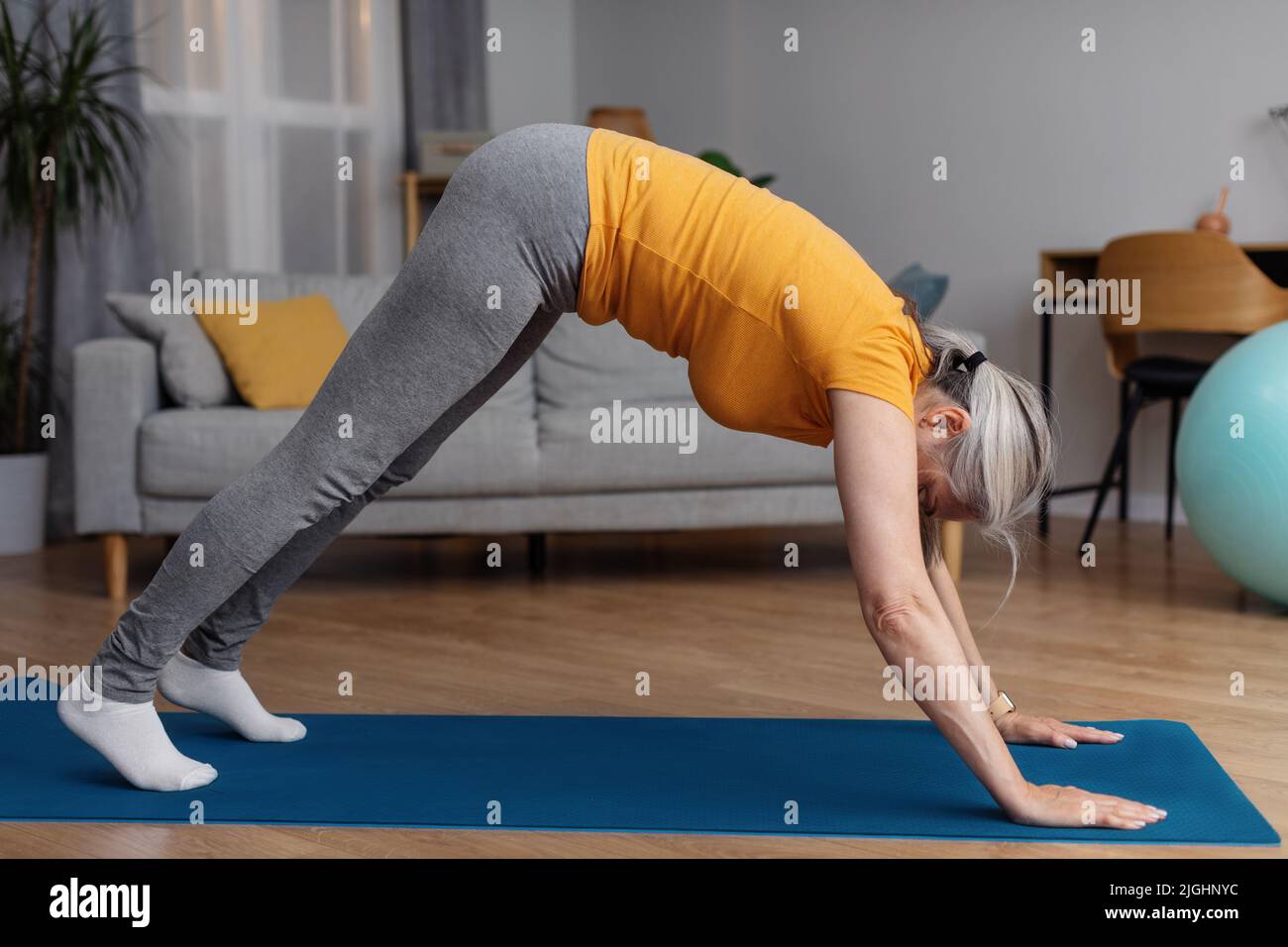 Inländische Yoga-Praxis. Flexible ältere Frau in nach unten schauenden Hund Asana stehend, Training auf Fitness-Matte zu Hause Stockfoto