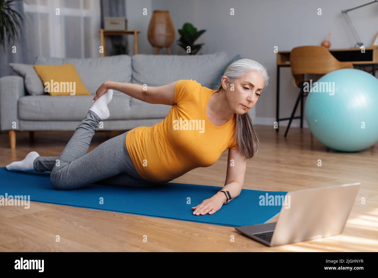 Häusliche Ausbildung. Aktive ältere Frau tut Beine Quad Stretching-Übungen, während Video-Tutorial auf Laptop Stockfoto