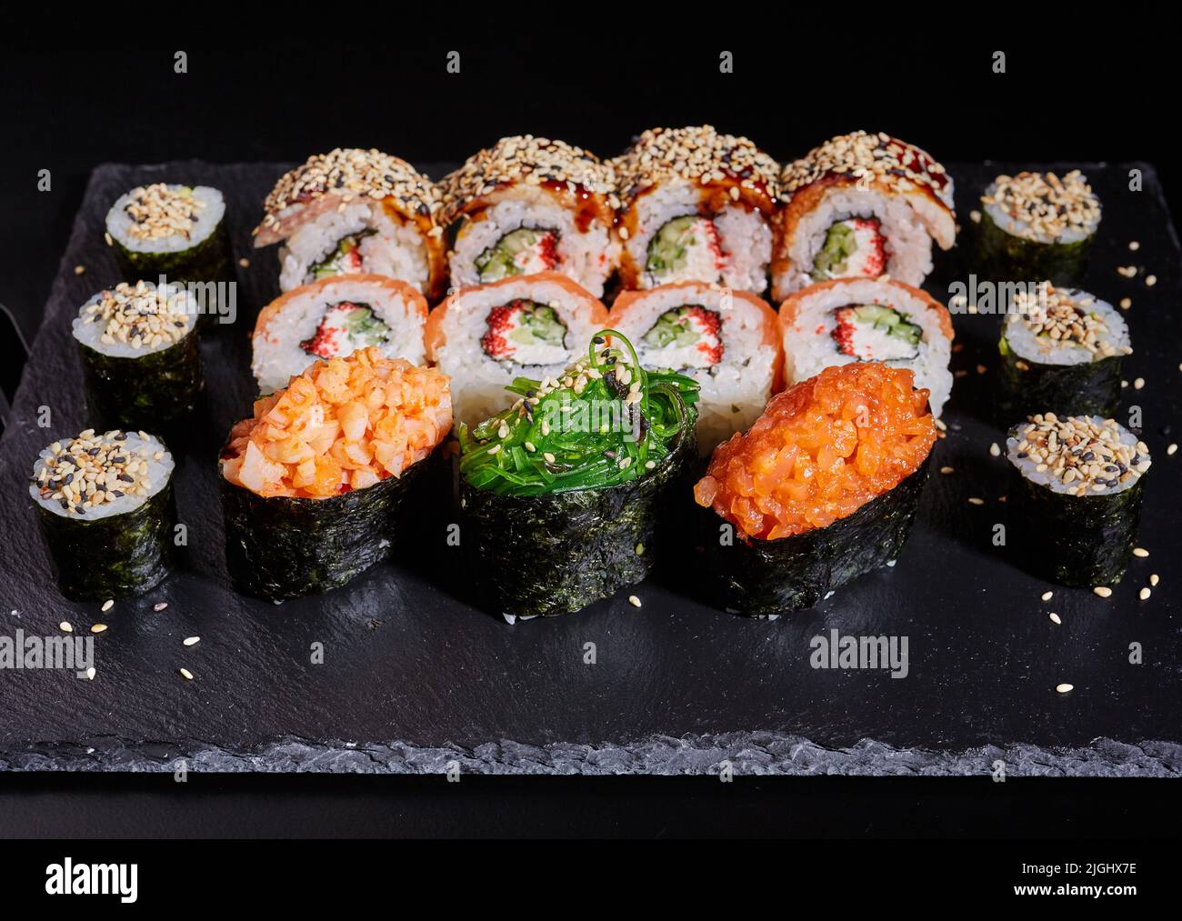 Nahaufnahme eines Sushi-Sets auf einem schwarzen Steinbrett auf einem dunklen Tisch. Nahaufnahme verschiedener Sushi-Rollen, die mit Sesamsamen auf einem schwarzen Stein bestreut sind Stockfoto