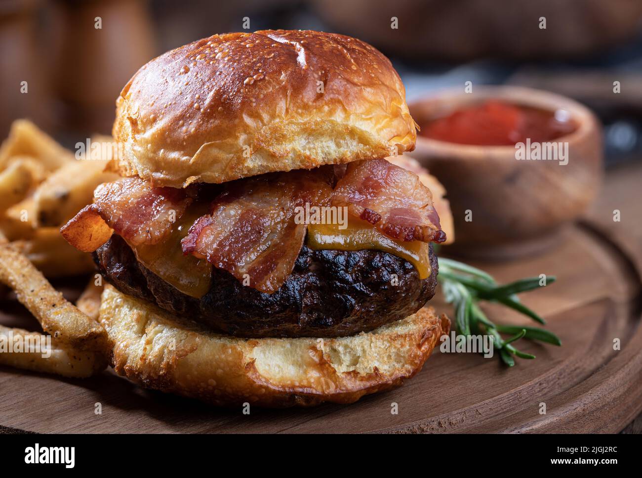 Nahaufnahme von Speck-Cheeseburger auf getoastetem Brötchen mit pommes auf einer Holzplatte Stockfoto