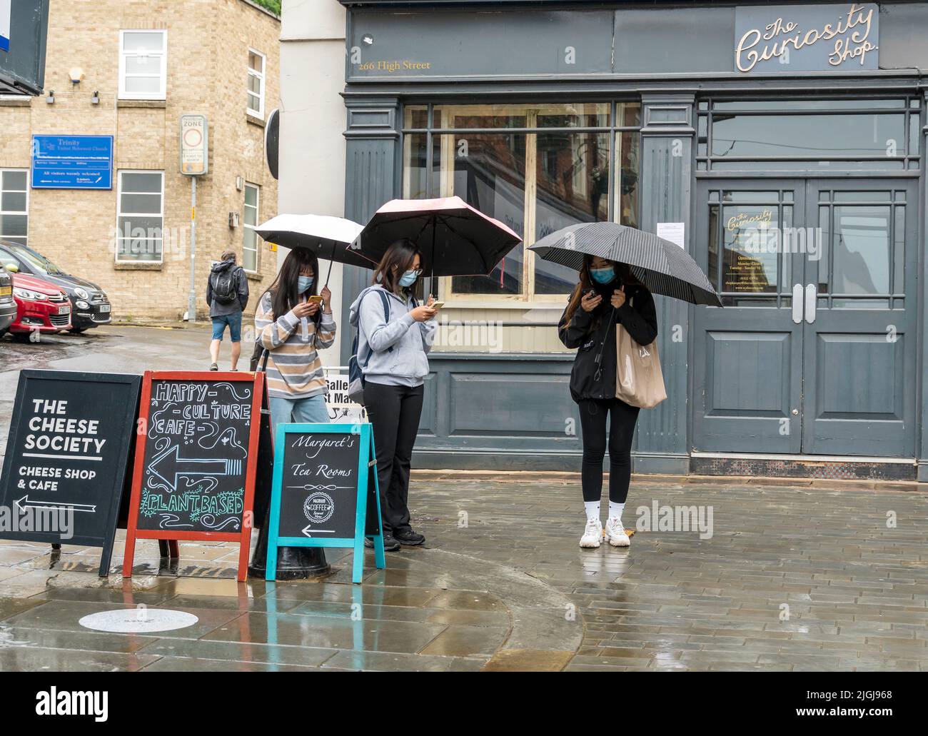 Drei junge orientalische Damen mit offenen Regenschirmen in Regenguss überprüfen Handys für Richtungen High Street Lincoln City 2022 Stockfoto