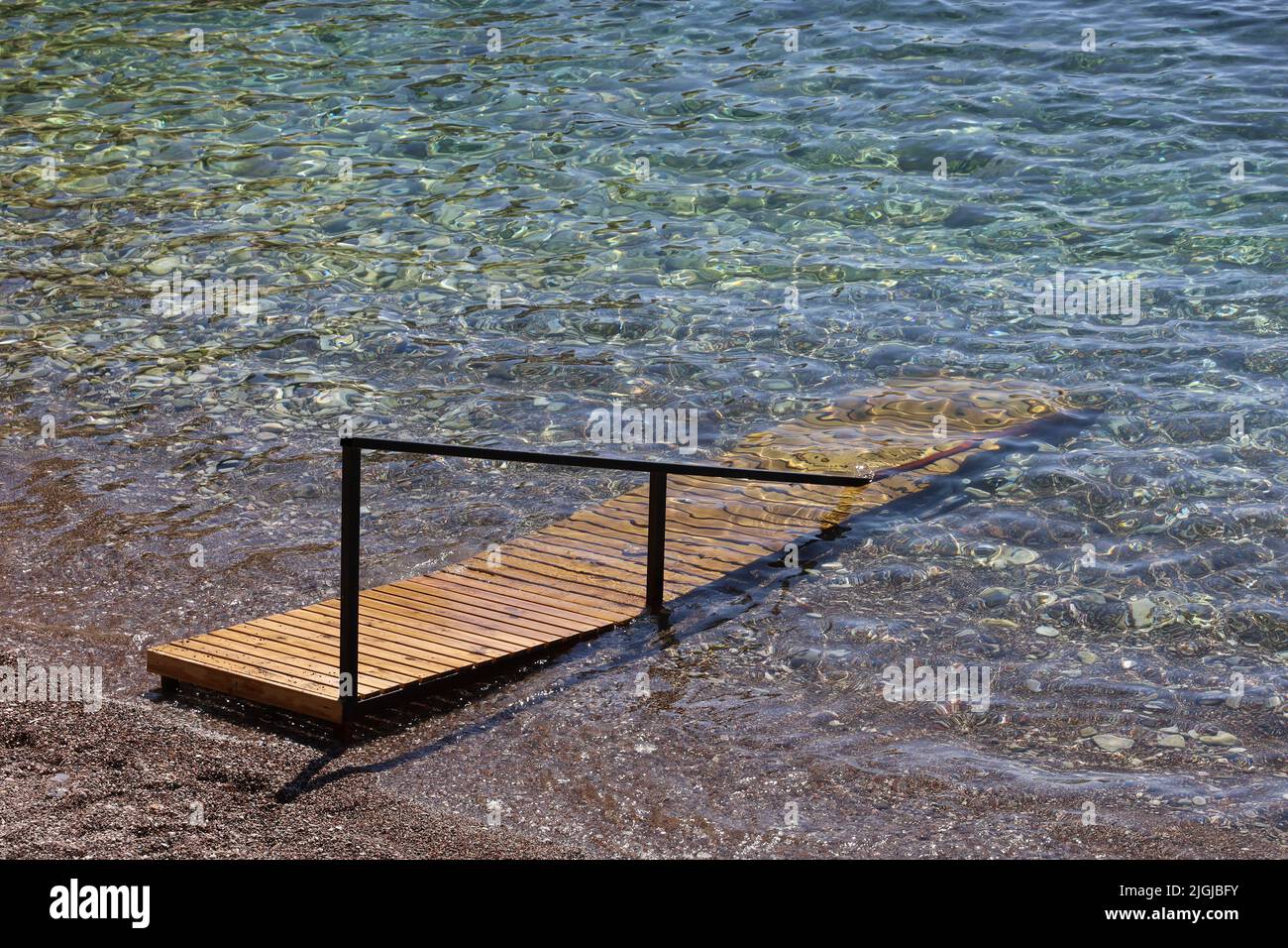 Leerer Kiesstrand mit Holzrampe, die in das transparente Wasser führt. Urlaub im Sommerresort Stockfoto