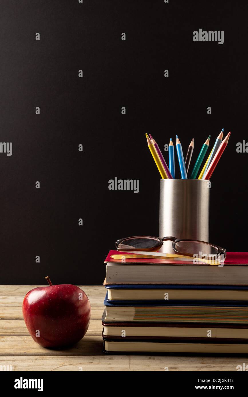 Vertikale Abbildung von Büchern, Gläsern, Buntstiften im Behälter und Apfel auf schwarzer Oberfläche Stockfoto