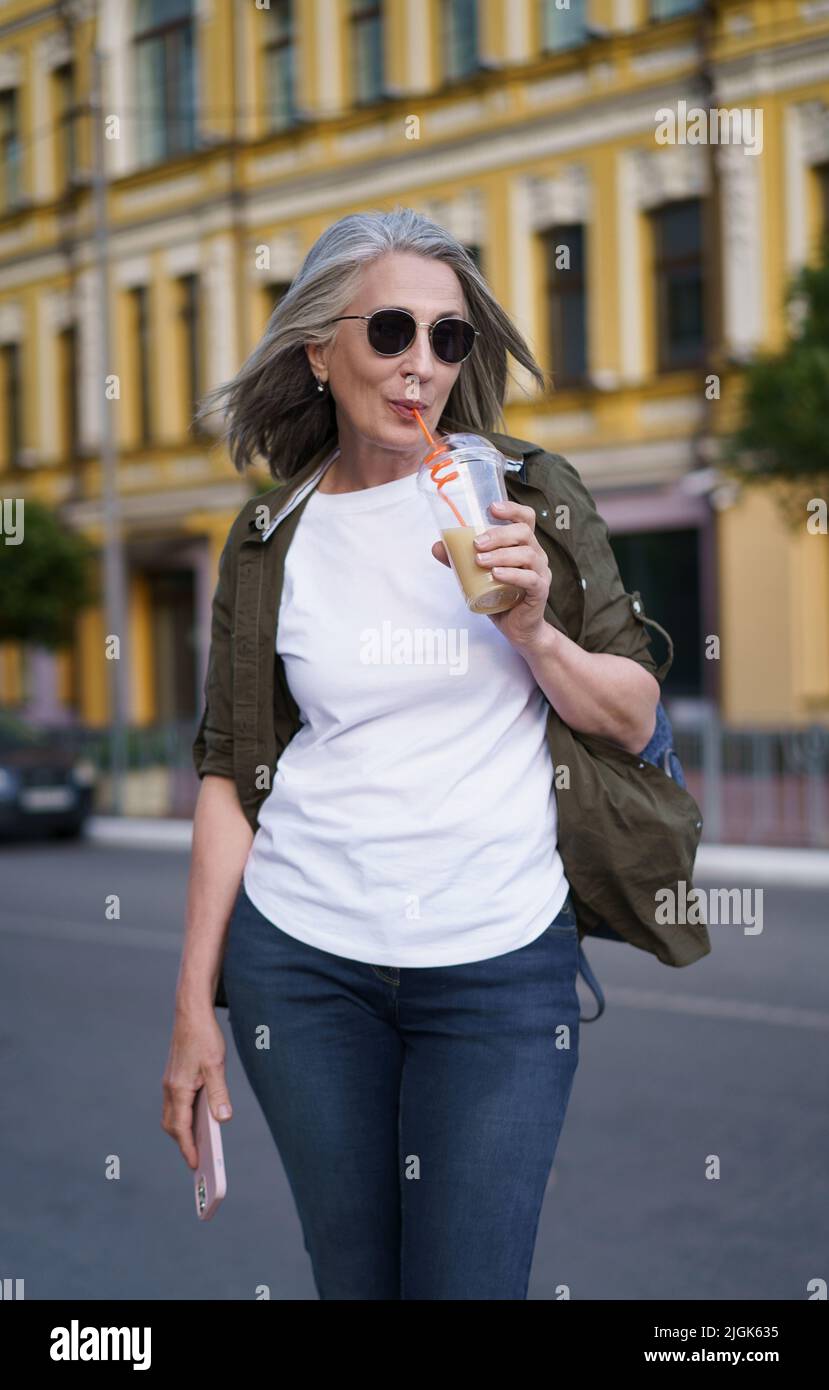 Ältere europäische Frau mit grauem Haar glücklich genießen Freizeit Reisen mit Telefon halten, während Saft auf dem Weg mit Plastikbecher im Hintergrund der Stadt. Genießen Sie das Leben reife Frau. Stockfoto