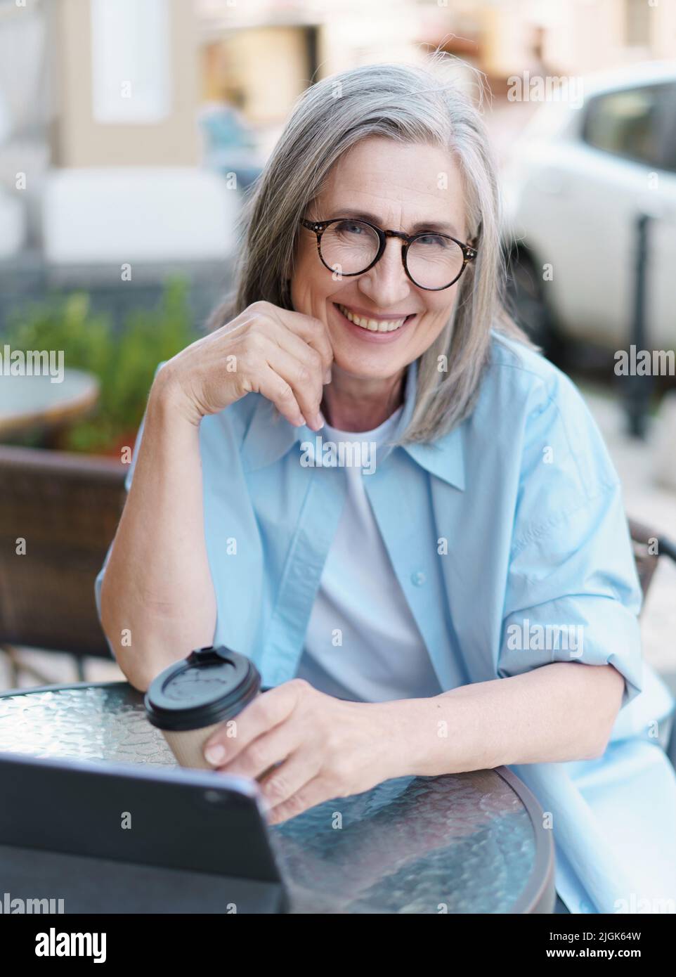 Europäische grauhaarige reife Frau trinkt Kaffee, während sie mit einem digitalen Tablet im kleinen Straßencafé im Internet surft. Reife Frau, die im Stadtcafe arbeitet und weißes T-Shirt und blaues Hemd trägt. Stockfoto