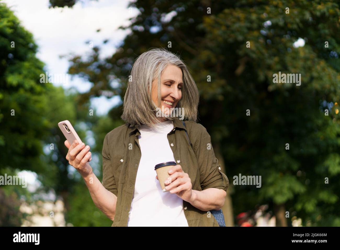 Reife europäische Frau mit grauem Haar freut sich nach der Arbeit oder auf Reisen mit dem Telefon, während sie unterwegs mit einer Papiertasse im Stadtgarten oder Park Kaffee trinkt. Genießen Sie das Leben reife Frau. Stockfoto