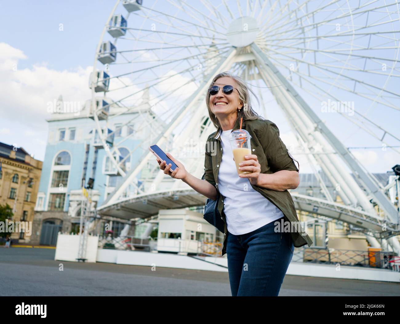 Europäische charmante grauhaarige Frau glücklich genießen Freizeit nach der Arbeit oder auf Reisen, Telefon halten, während Saft auf dem Weg mit Plastikbecher in der Stadt und Riesenrad auf dem Hintergrund. Stockfoto
