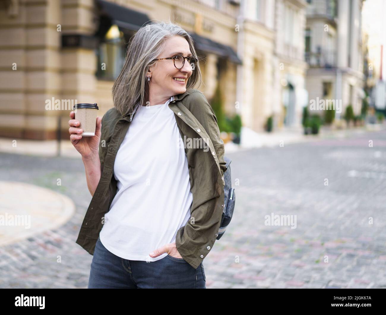 Geschäftsfrau halten Einweg-Tasse mit heißem Kaffee. Die reife Frau verbringt ihre Freizeit in der europäischen Altstadt und genießt eine Tasse Tee und Kaffee. Grauhaarige Frau in Casual mit Rucksack. Stockfoto