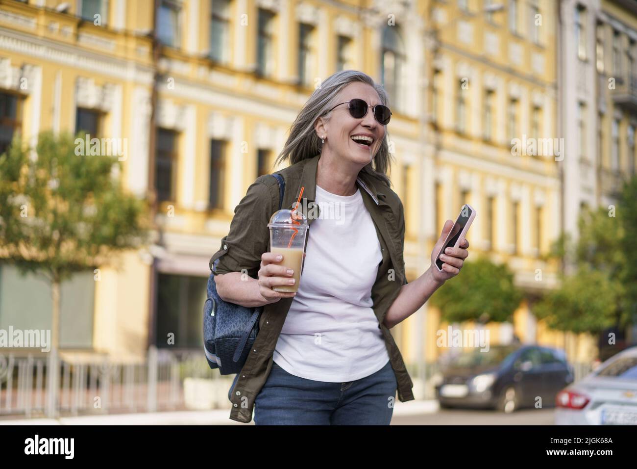 Europäische ältere Frau mit grauen Haaren glücklich genießen Freizeit nach der Arbeit oder auf Reisen mit Telefon halten, während Saft auf dem Weg mit Plastikbecher im Hintergrund der Stadt. Genießen Sie das Leben reife Frau. Stockfoto