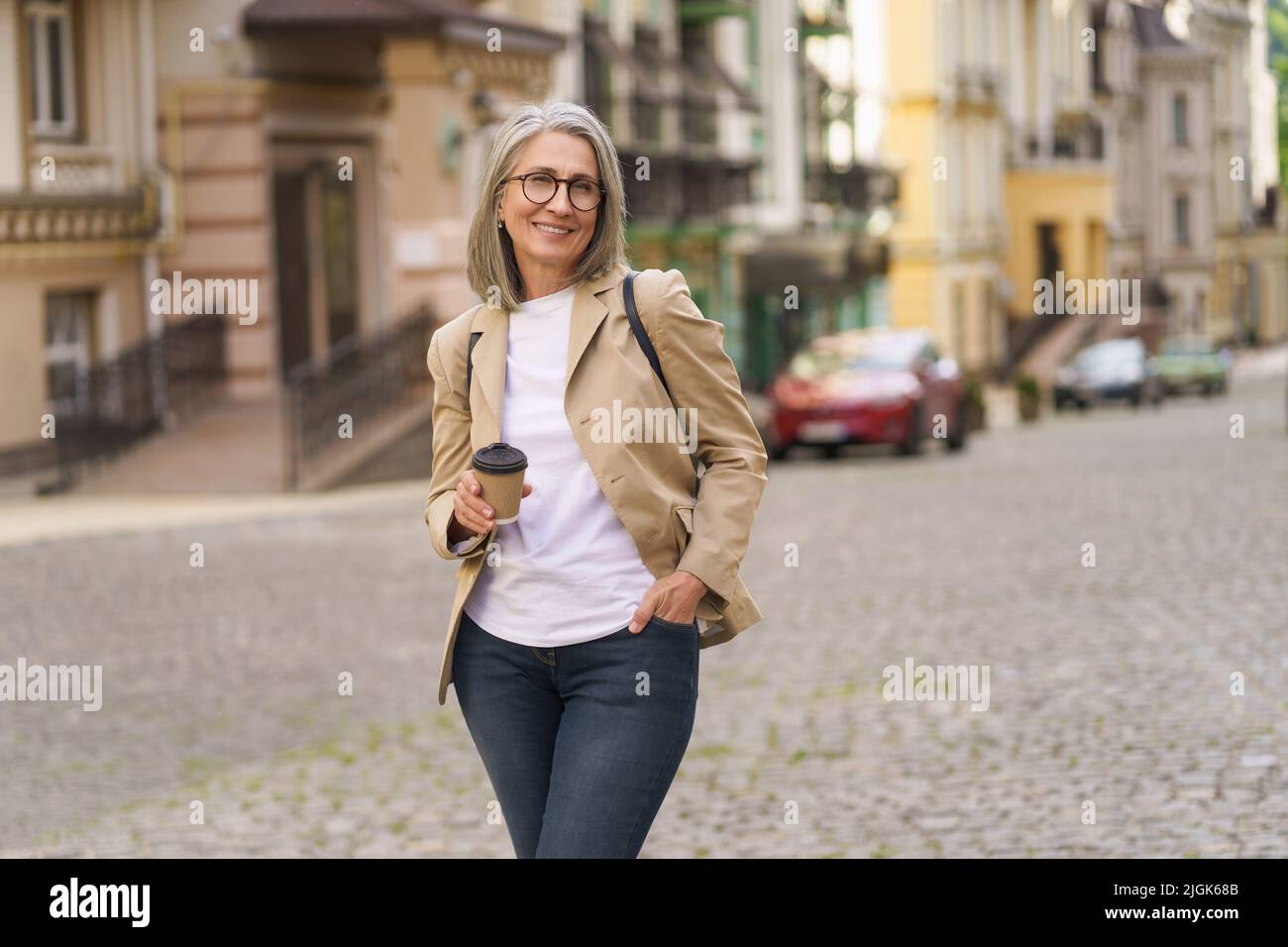 Reife Geschäftsfrau in lässig formell halten Einweg-Tasse mit heißem Kaffee steht im Freien der Altstadt. Die reife Frau verbringt ihre Freizeit in der europäischen Stadt und genießt eine Tasse Tee und Kaffee. Stockfoto