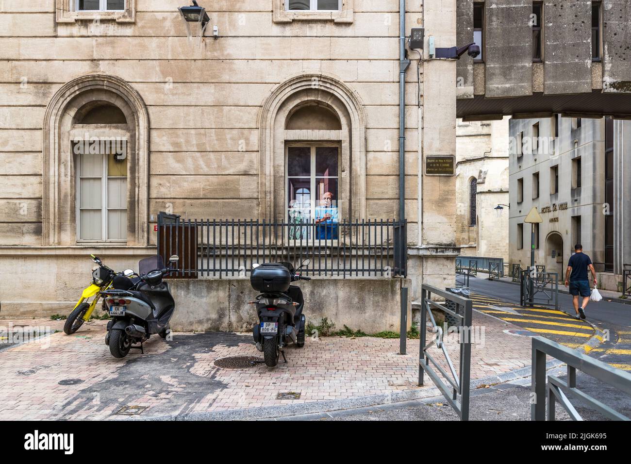Ein Mann blickt aus dem Fenster einer historischen Sandsteinfassade in der Altstadt von Avignon, Frankreich; zwei Motorroller sind unten geparkt, mit dem Rathaus (Hôtel de Ville) im Hintergrund. Stockfoto