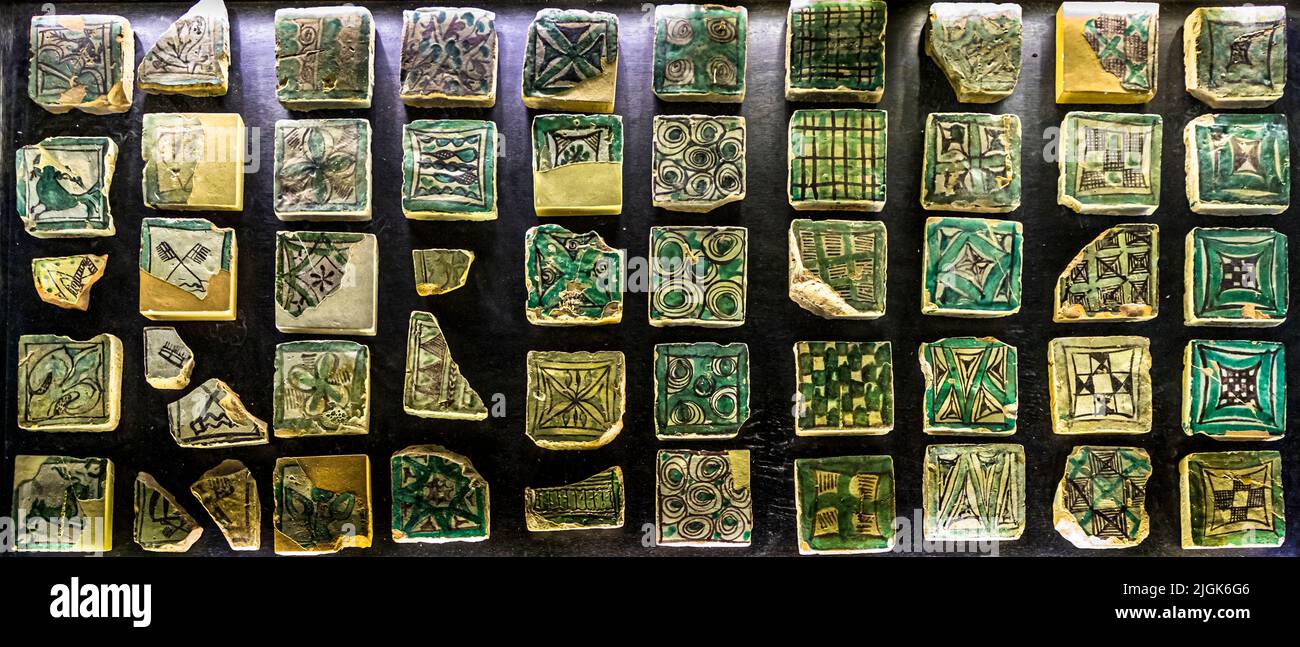 Kleine mittelalterliche Bodenziegel mit geometrischen Mustern – Kreise, Rauten, Gitterlinien – liegen dicht gepackt in einer Vitrine im Papstpalast von Avignon, Frankreich. Stockfoto