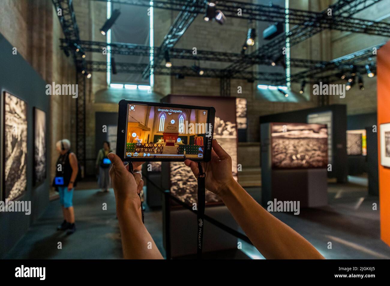 Ein Histopad-Tablet rekonstruiert die "Grande Chapelle" des Papstpalastes von Avignon, Frankreich. Im Hintergrund hängen großformatige Schwarz-Weiß-Fotografien aus Sebastião Salgados Ausstellung "Amazônia" unter Beleuchtungsgerüsten. Stockfoto