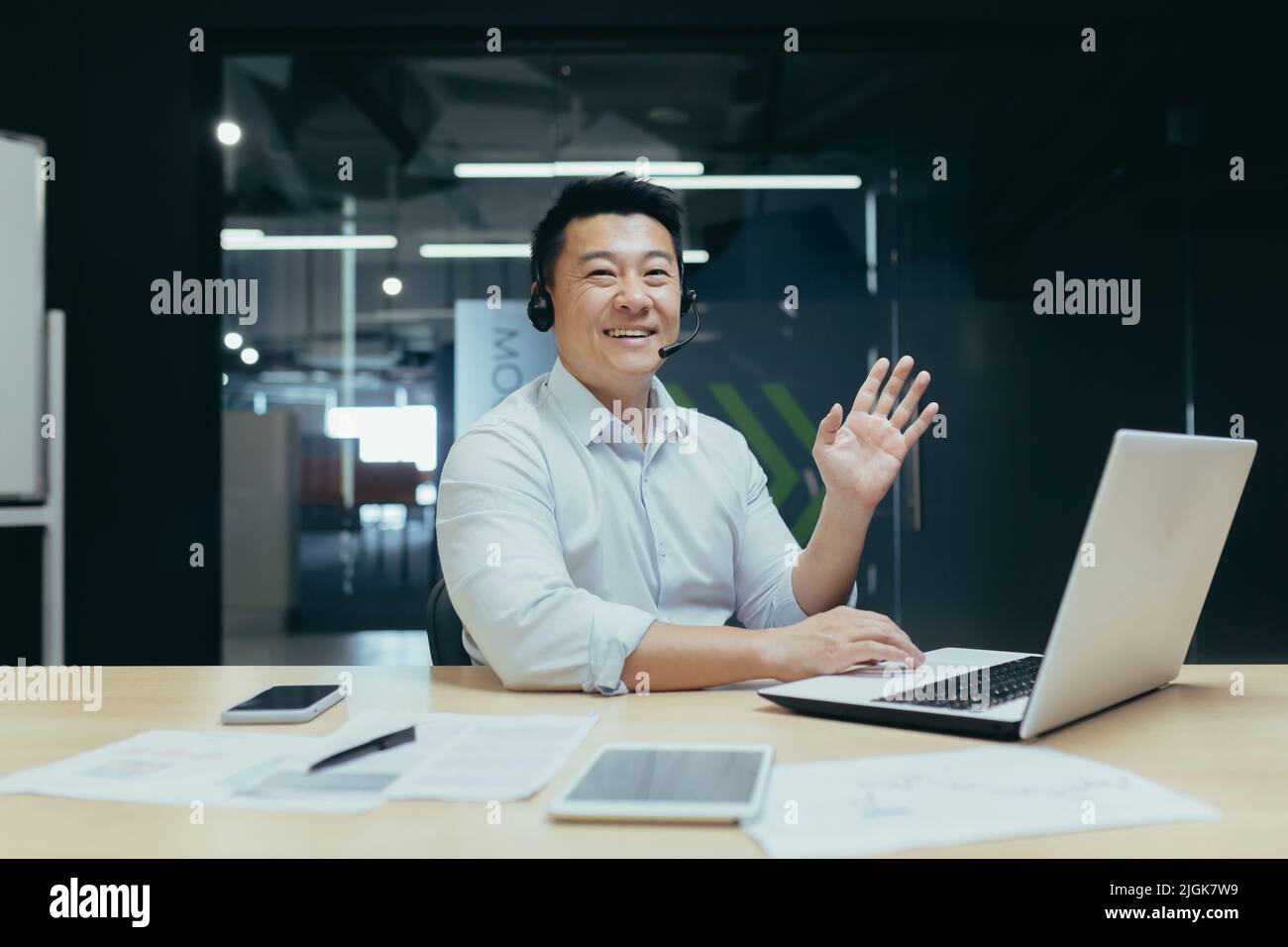 Porträt eines asiatischen Geschäftsmannes, der die Kamera anschaut und eine winkende Grußgeste zeigt, eines Mannes mit Headset für Videoanruf Stockfoto