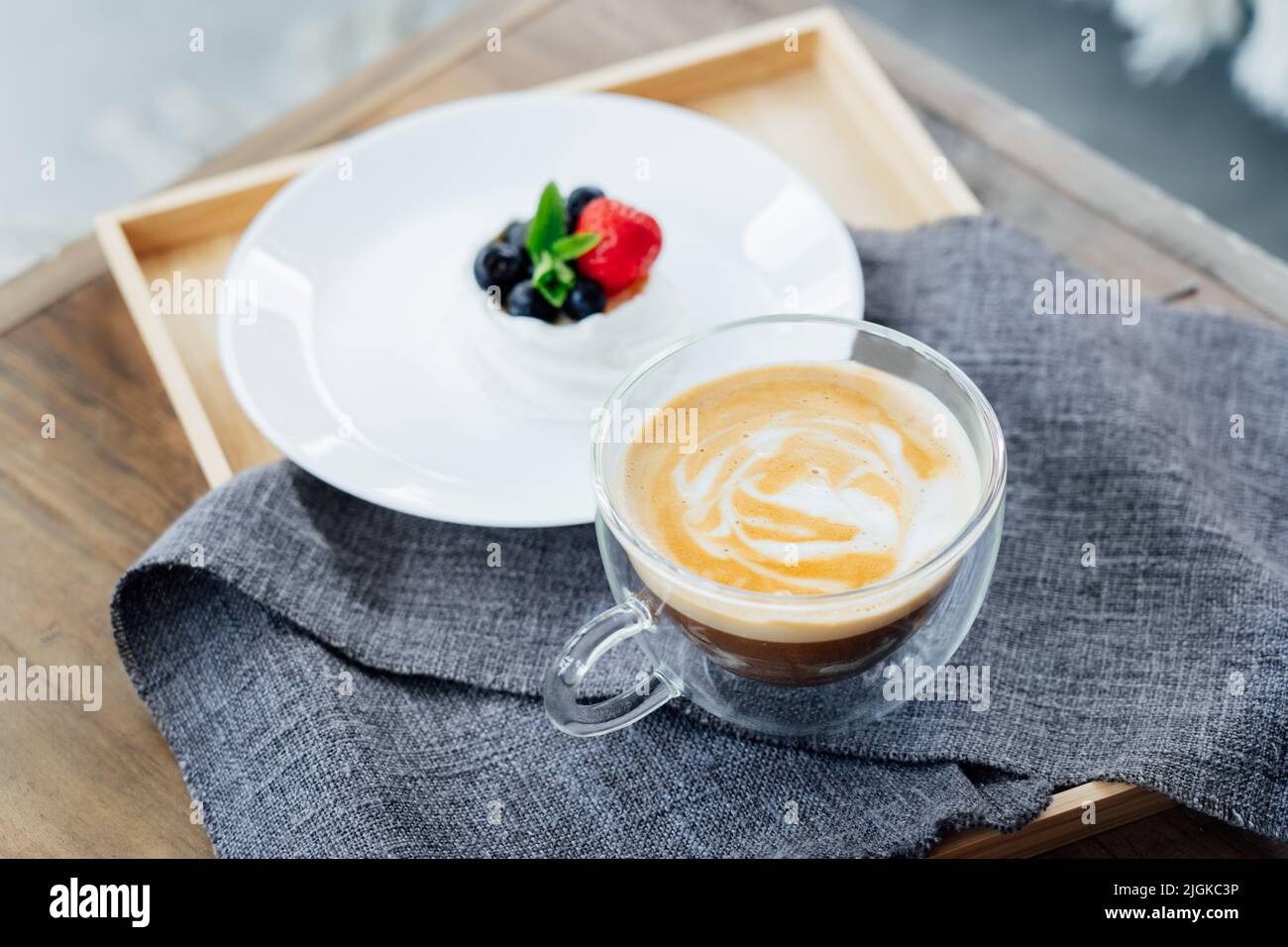 Pavlova Dessert Mini-Kuchennest und eine Tasse Latte-Kaffee auf dem Holztablett mit grauer Serviette auf dem Tisch. Kaffeezeit. Um eine Pause zu machen und sich zu entspannen. Sel Stockfoto