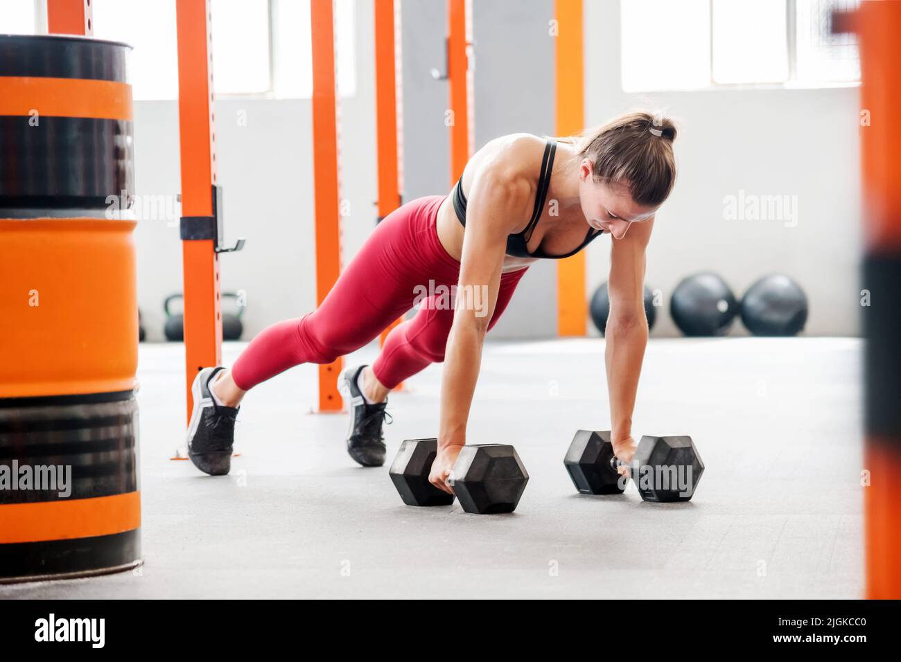 Starke Sportlerin in Activewear mit Pferdeschwanz macht Plank mit Kurzhanteln beim Funktionstraining in geräumiger leichter Turnhalle Stockfoto
