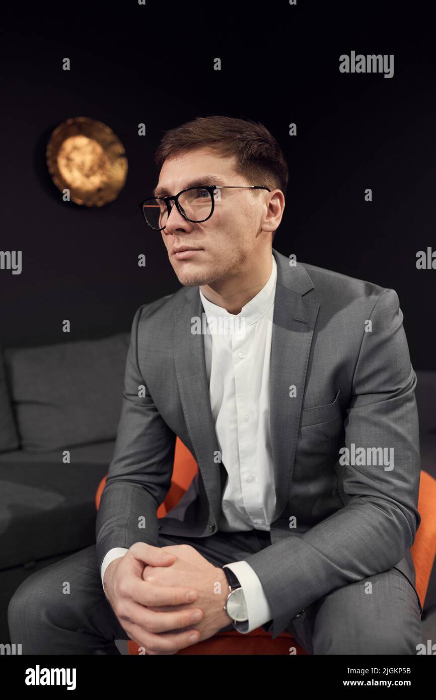 Seriöser junger Geschäftsmann in Anzug und Brille sitzt auf einem bequemen Stuhl und spricht beim Interview mit einer wichtigen Person Stockfoto