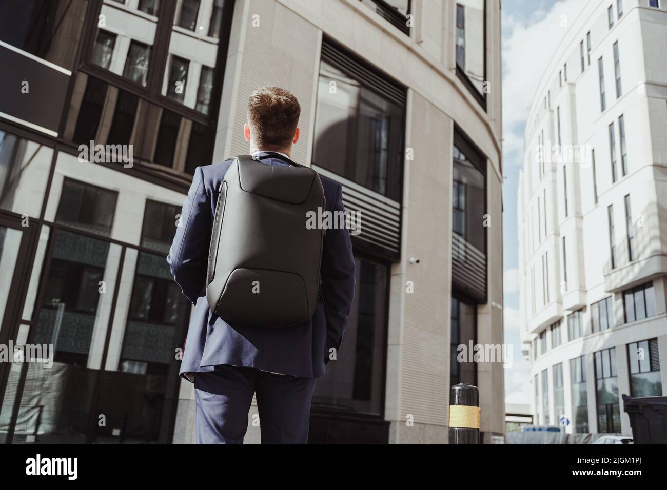 Der junge Angestellte in einem Anzug mit Rucksack steht vor einem Firmengebäude und schaut nach oben, von hinten. Praktikum, Vorstellungsgespräch, neuer Job Stockfoto