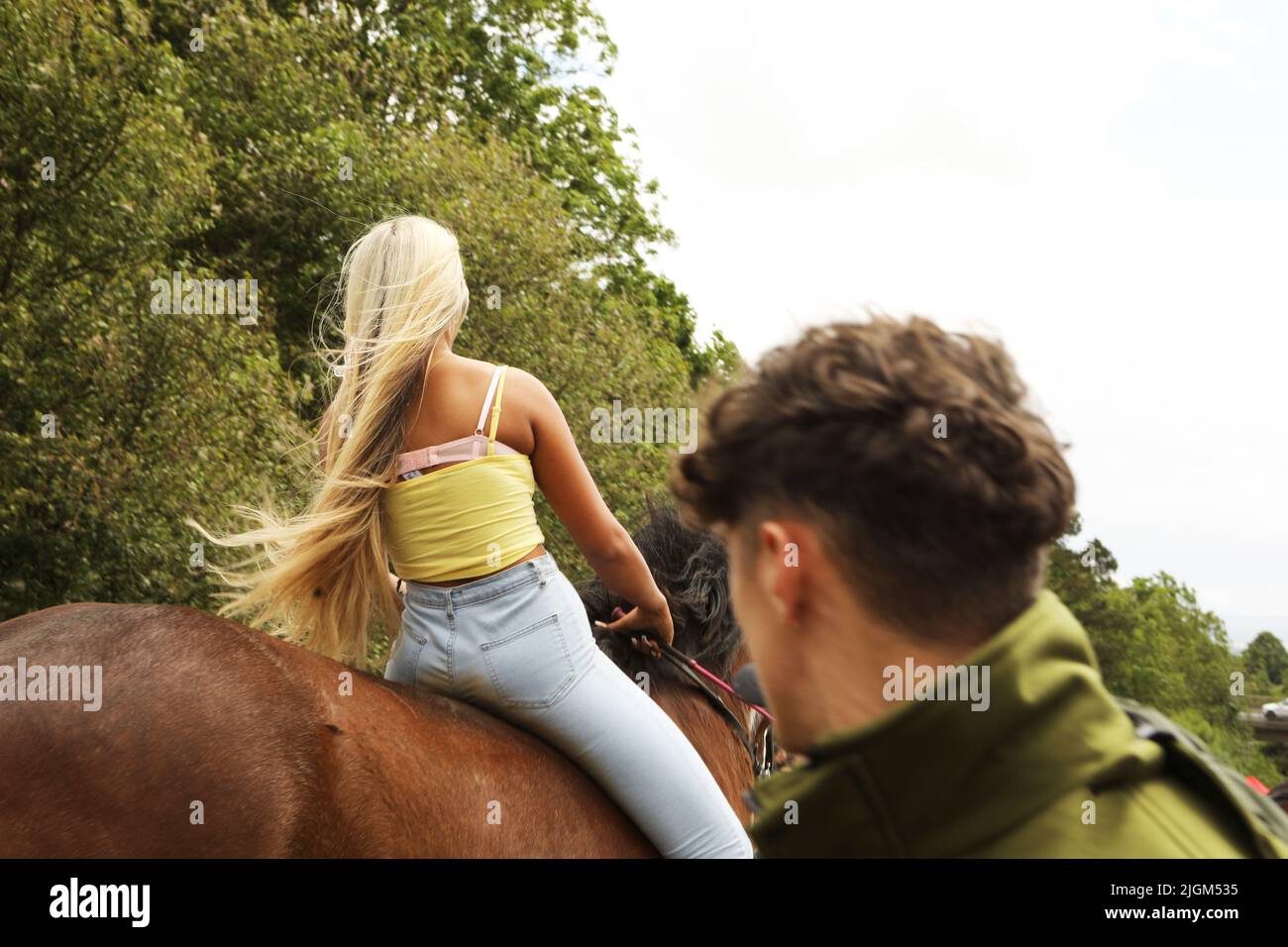 Ein junger Mann dreht seinen Kopf und beobachtet eine junge Frau mit langen blonden Haaren, wie sie auf einem Shire-Pferd auf der Straße reitet. Appleby Horse Fair Stockfoto