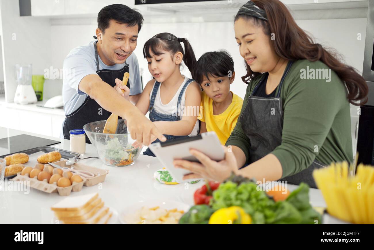 Glückliche asiatische Eltern und ihre Kinder haben Spaß beim Kochen und nach Rezept im Internet. Stockfoto