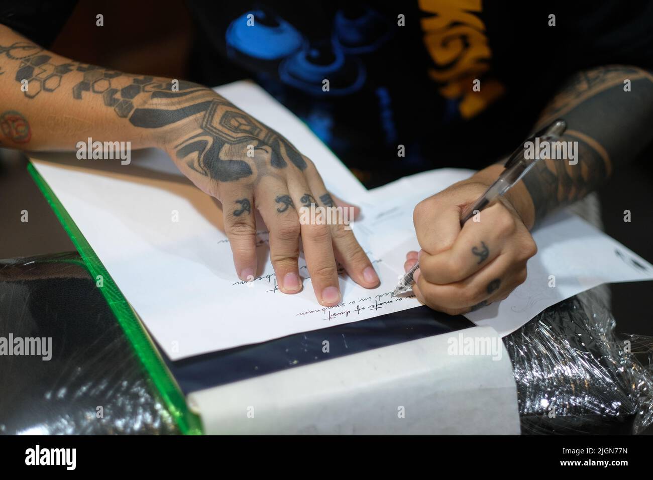 Professioneller Tattoo-Künstler, Arme gefüllt mit grafischen Designs, Schablonen kursive Schrift auf weißem Papier mit einem Stift zur Vorbereitung auf eine Tattoo-Sitzung. Stockfoto