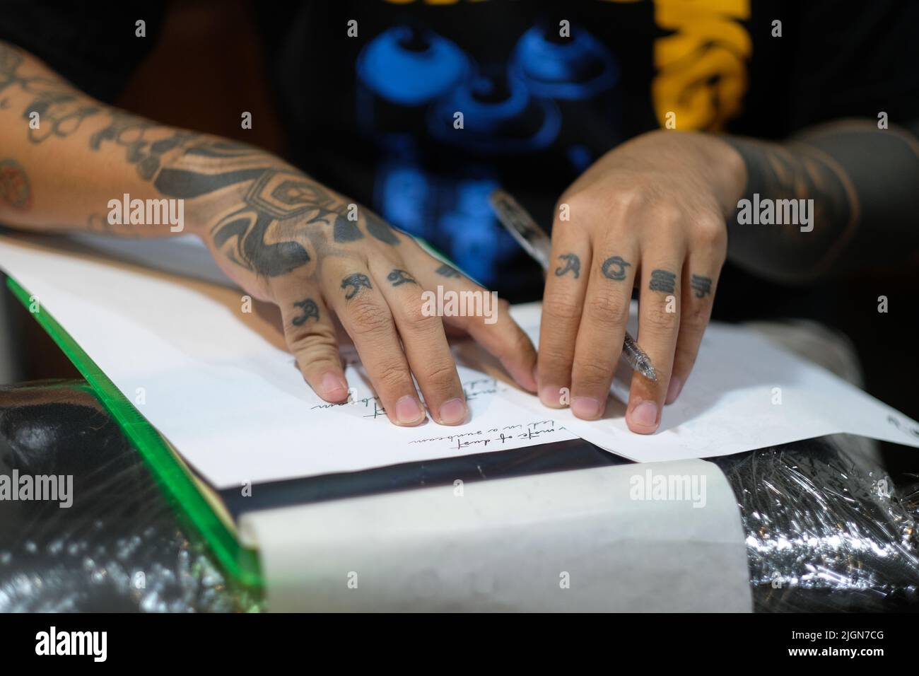 Nahaufnahme der eingekeilten Finger des Tätowierers, während er pausiert, während er kursive Schrift mit einem Stift auf weißes Papier schminkt, um sich auf eine Tätowierungssitzung vorzubereiten. Stockfoto