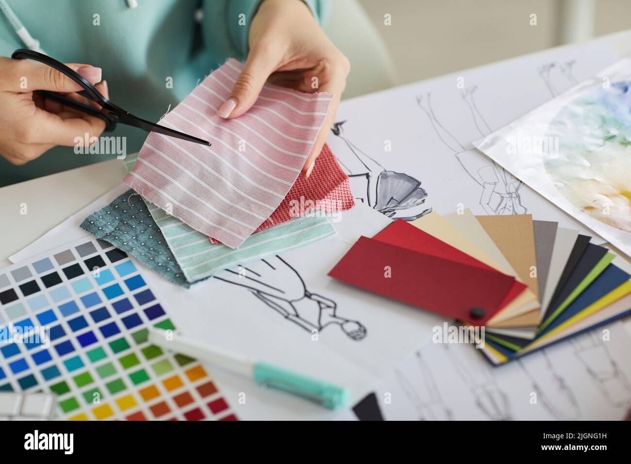 Nahaufnahme einer nicht erkennbaren Frau, die mit Paletten und Modezeichnungen am Schreibtisch sitzt und im Schneideratelier Stoff schneidet Stockfoto