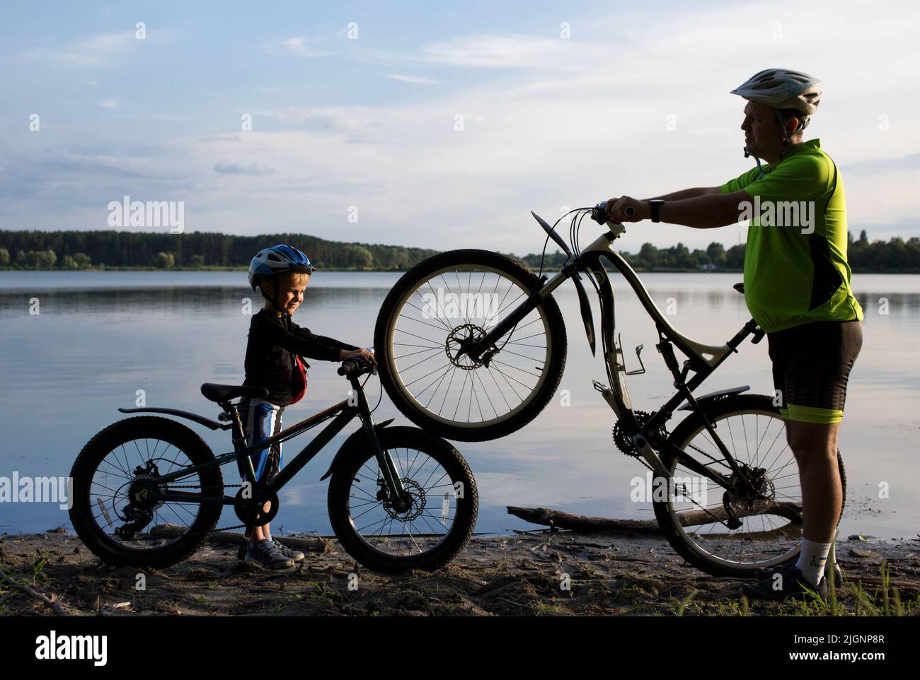 Silhouetten eines erwachsenen Mannes und eines Jungen mit zwei Fahrrädern in Helmen in der Nähe des Sees bei Sonnenuntergang. Radfahren, gesunder Lebensstil. Sei wie Papa. Familienvertreib, d Stockfoto