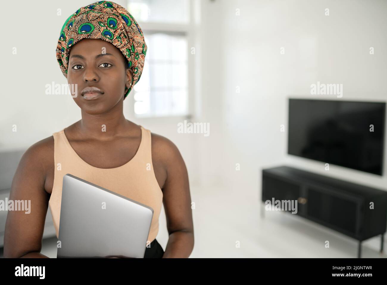 Porträt einer schönen afrikanischen Frau, die in einem modernen Zuhause mit einem Laptop in der Hand in die Kamera schaut und einen traditionellen Schal mit Kopfband trägt Stockfoto