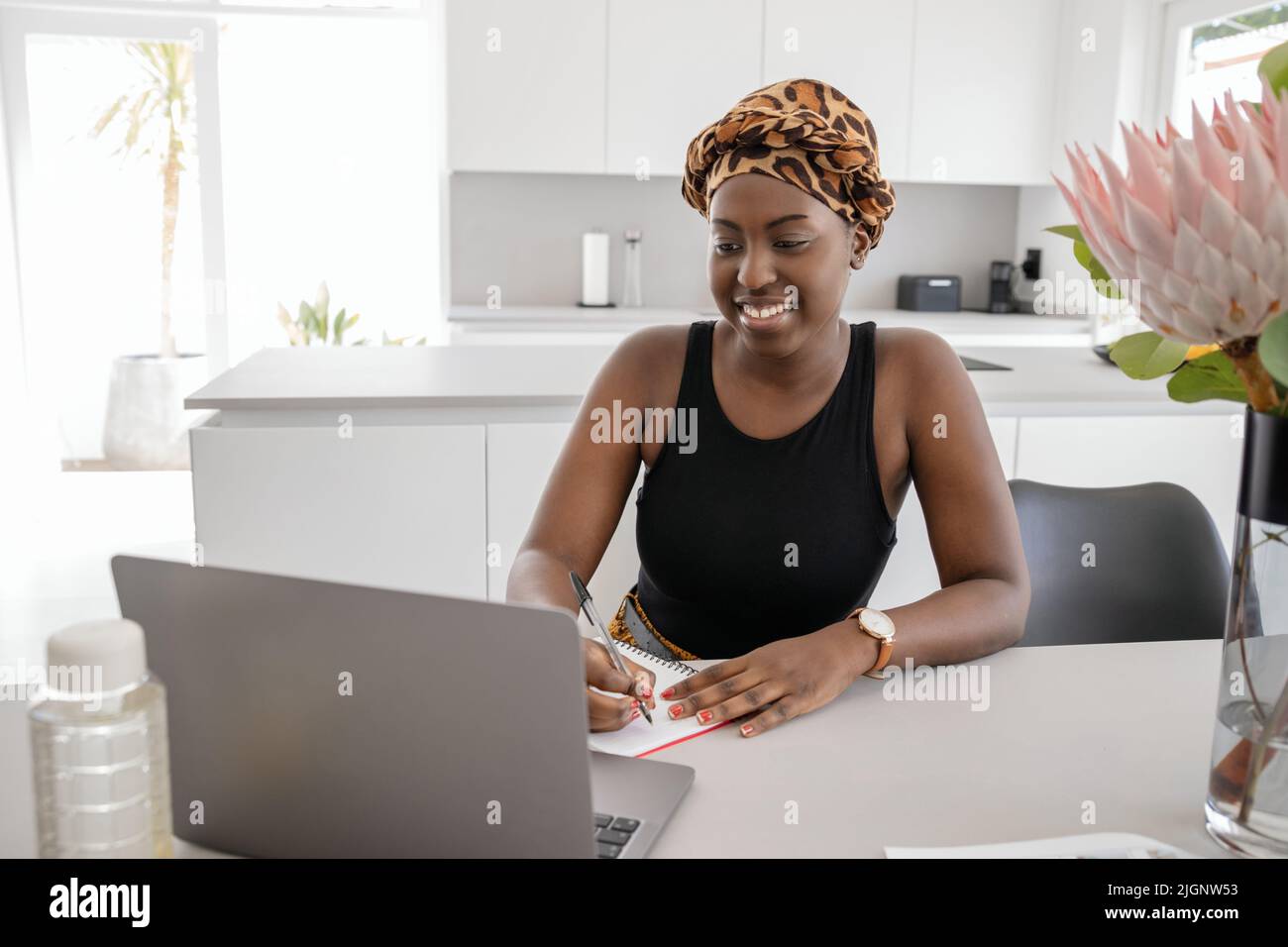 Afrikanische Frau, die von zu Hause aus studiert, auf einen Laptop schaut und Notizen schreibt. Laptop und Smartphone auf dem Schreibtisch des Heimbüros Stockfoto