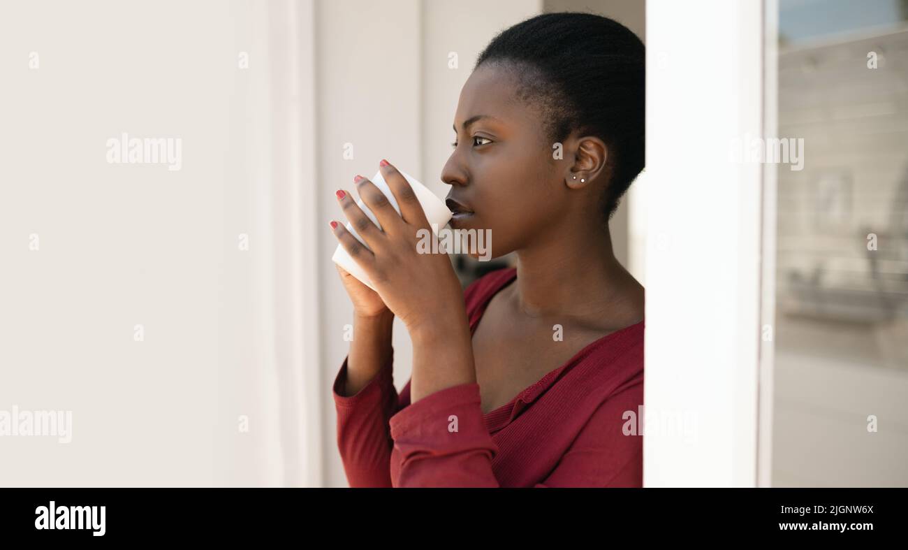 Porträt einer schönen afrikanischen Frau, die auf dem Weg zu Hause vor der Tür steht, einen Becher in der Hand hält und in die Ferne blickt Stockfoto